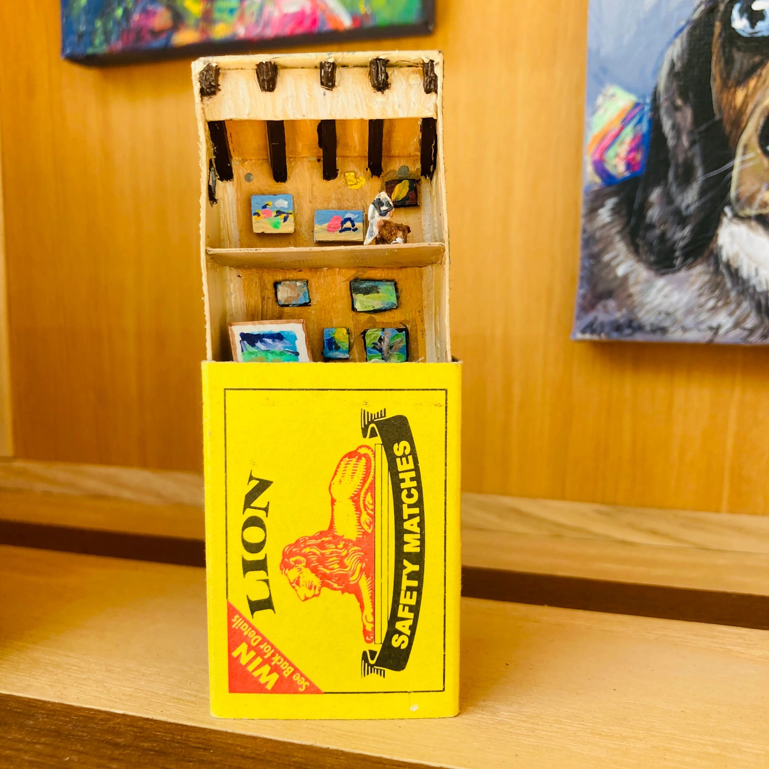 Miniature art gallery inside a matchbox with tiny paintings and a small figure.