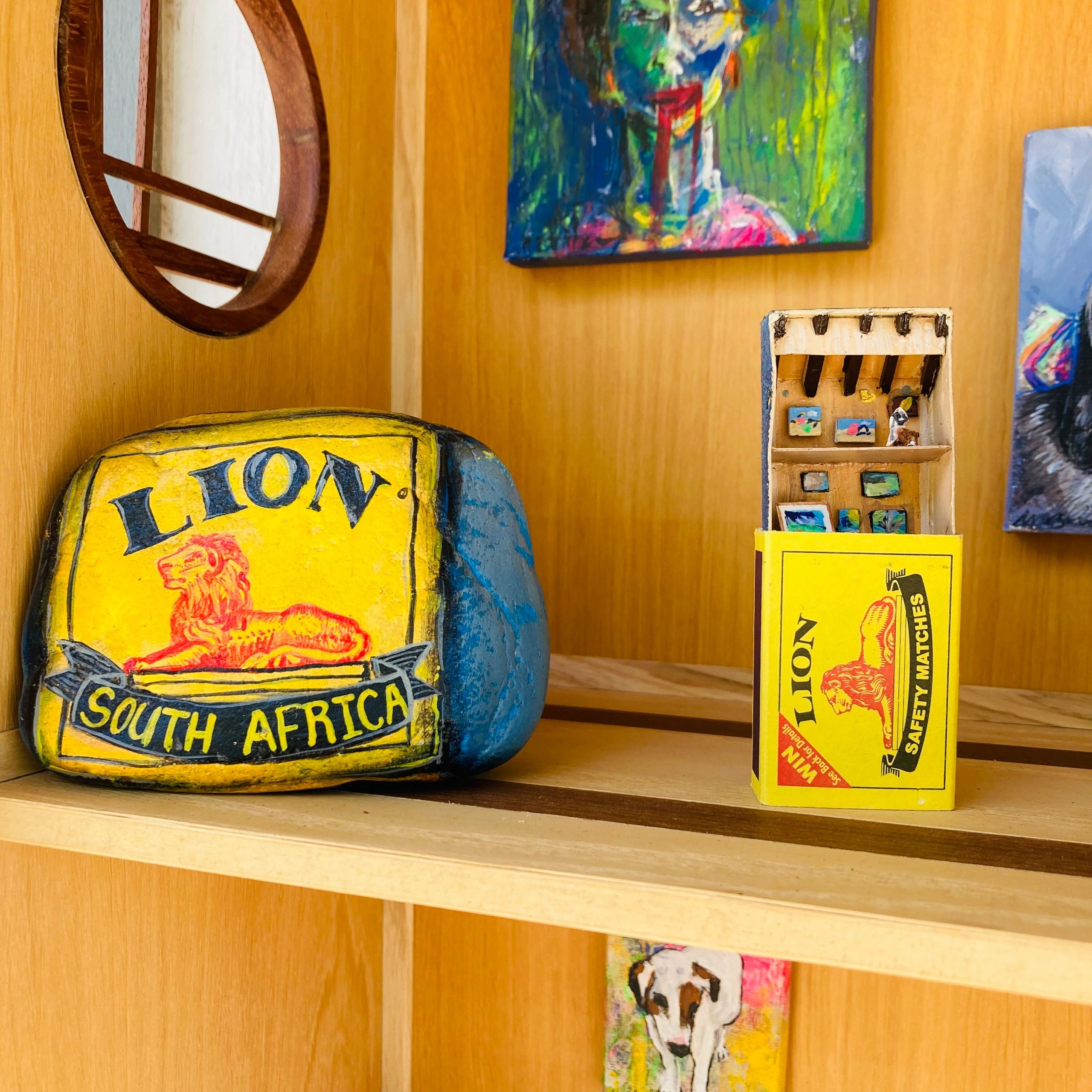 Vintage Lion South Africa matchbox and colorful paintings on a wooden shelf.