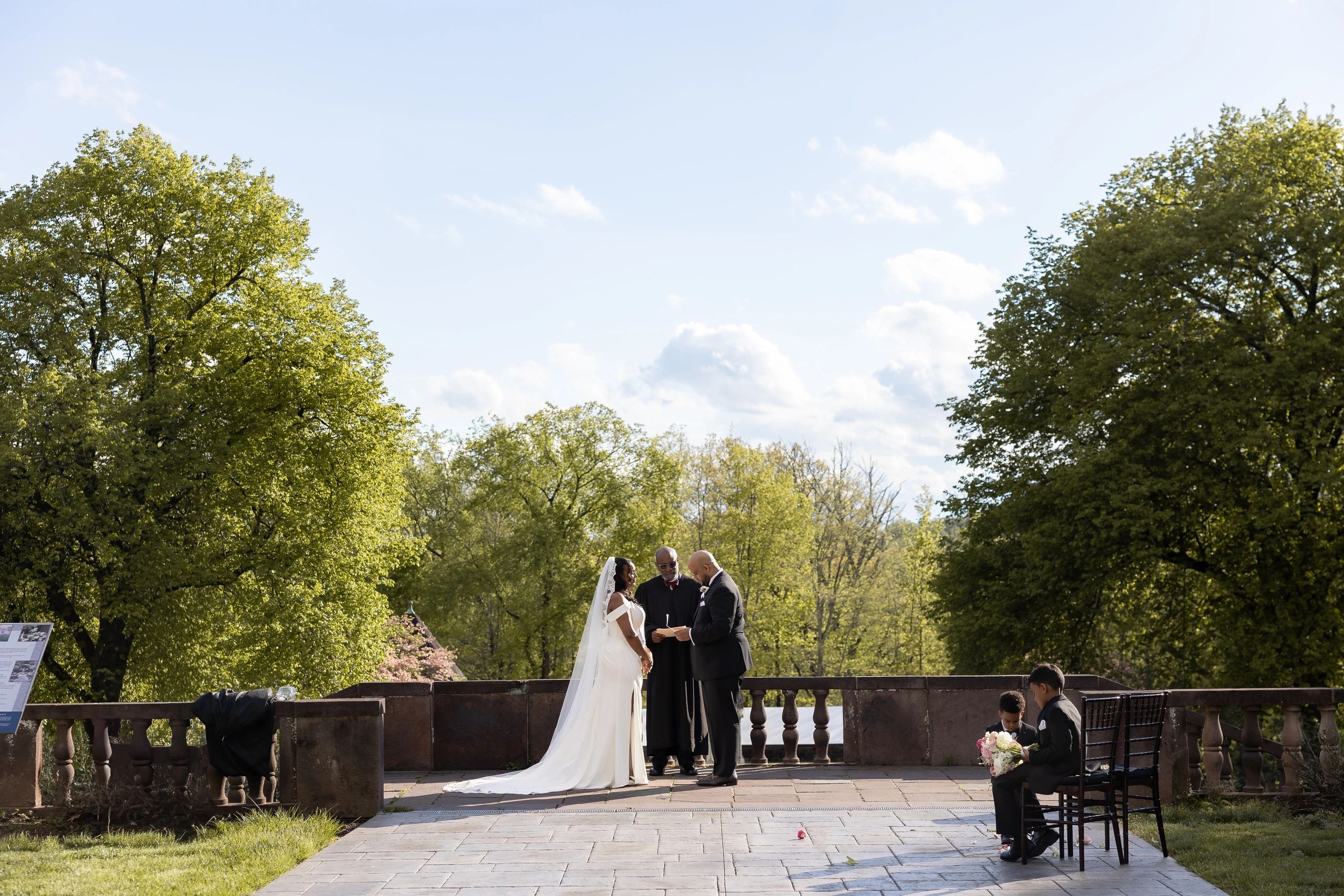 Outdoor wedding ceremony with bride, groom, and officiant surrounded by greenery.
