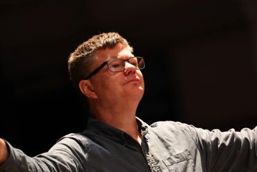 Man in glasses with arms outstretched, lit against a dark background.