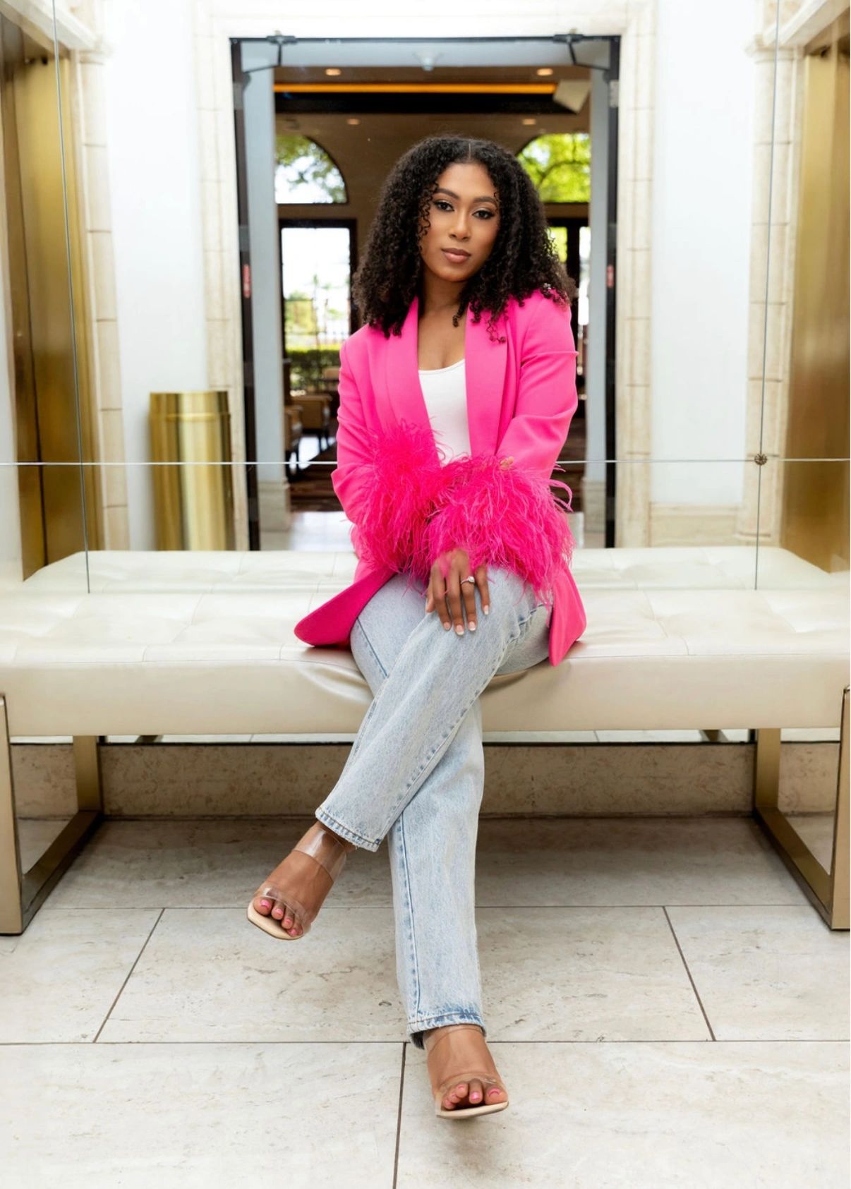 Woman seated on a bench wearing a pink feathered blazer and light jeans.