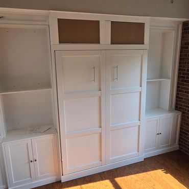 White built-in shelves and cabinets with a Murphy bed in a room with brick wall and wooden floor.