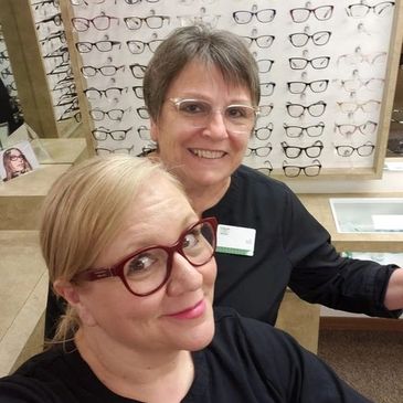 Two women wearing glasses and smiling in front of a glasses frame display