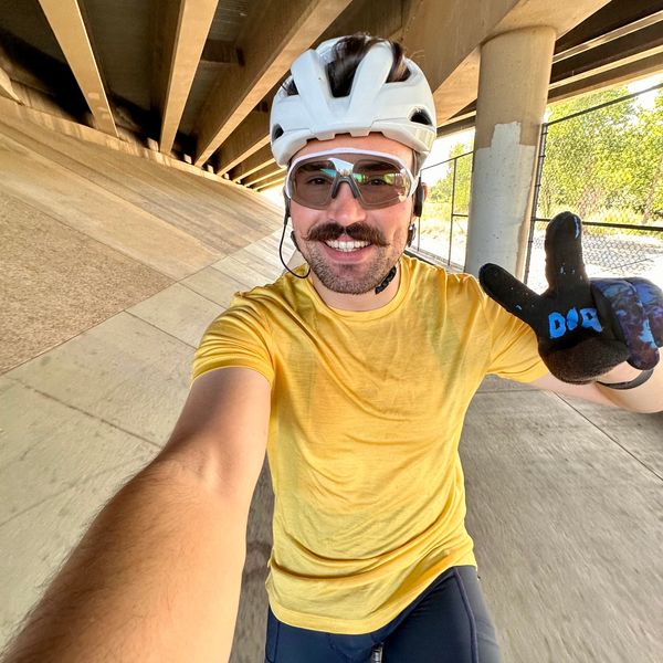 Smiling cyclist wearing a yellow shirt and helmet takes a selfie under a bridge.