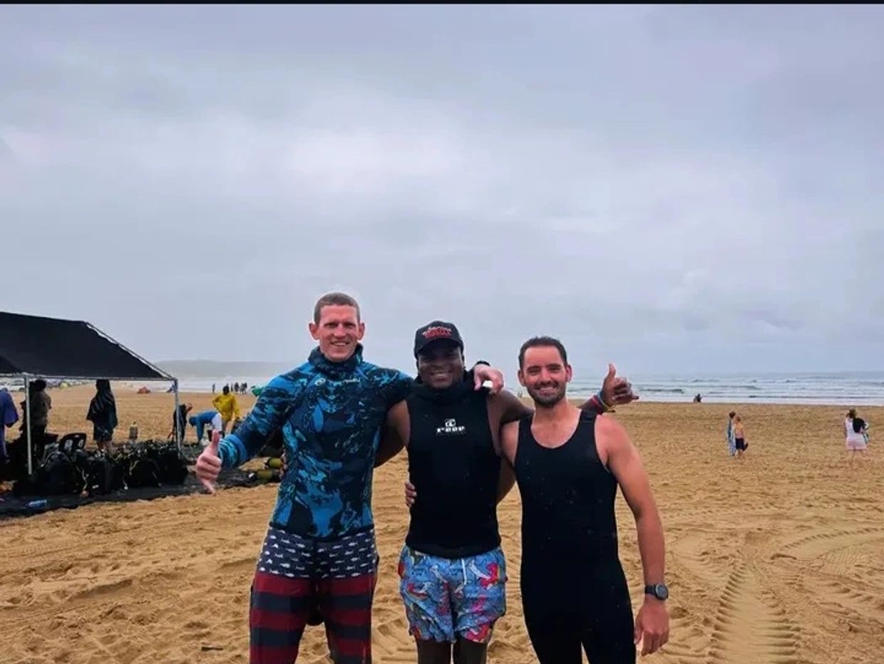 Three friends in wetsuits posing happily on a beach with thumbs up.