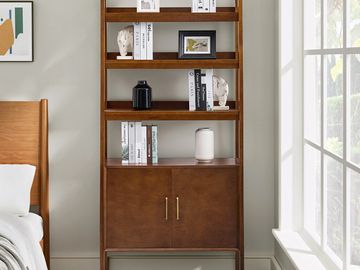 Wooden bookshelf with books, vases, and framed photo in a sunlit bedroom corner.