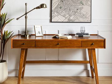 Mid-century wooden desk with brass lamp, framed picture, books, and plant.