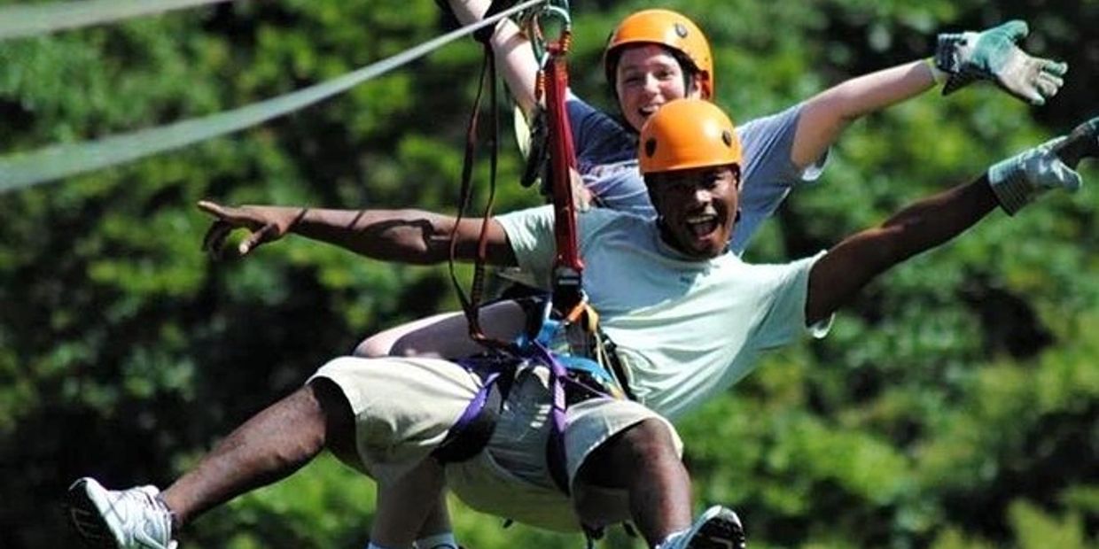 Two people ziplining with helmets and gloves, smiling.