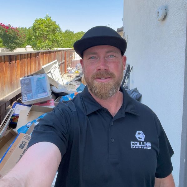 Man in black uniform smiling outdoors near a pile of boxes and trash.