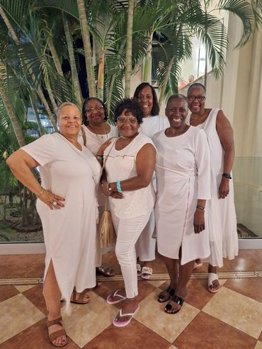 Six women dressed in white posing indoors near tropical plants, smiling warmly.