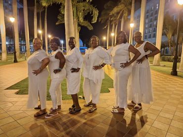 Six women dressed in white pose outdoors at night near palm trees and lampposts.
