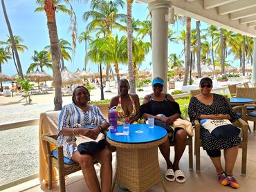 Four women sitting at a table by a tropical beach with palm trees and thatched umbrellas.