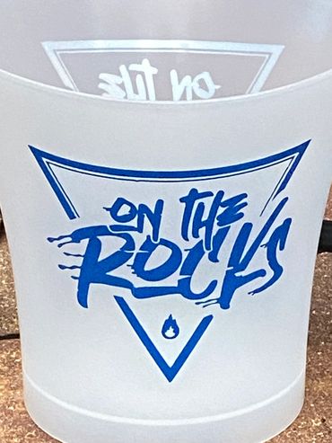 Frosted cup with blue 'On The Rocks' text on a brick surface.
