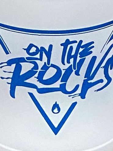 Frosted cup with blue 'On The Rocks' text on a brick surface.