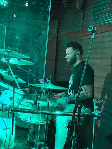 Drummer passionately playing behind a transparent shield on stage.