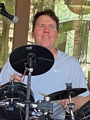 Man happily playing an electronic drum set indoors with a microphone nearby.