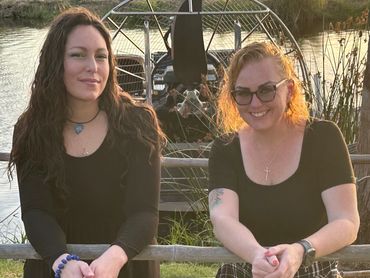 Two women in black tops smiling by a wooden fence near water and an airboat.