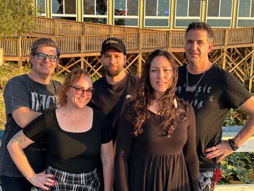 A group of five adults posing outdoors in warm sunlight near a wooden building.