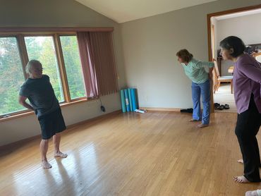 Three people stretching indoors on a wooden floor near a large window.