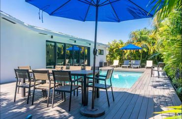 Outdoor pool area with dining table and blue umbrellas surrounded by lush greenery.