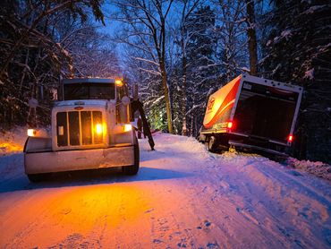 A snow-covered road with a stuck delivery truck and a tow truck at dusk.