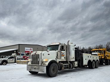 A white tow truck towing a yellow dump truck on a snowy lot.