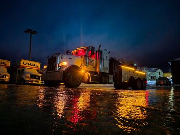 Illuminated tow truck at night reflecting on wet ground.