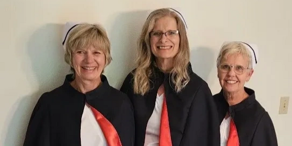 Three women in black capes with red lining and nurse caps smiling.