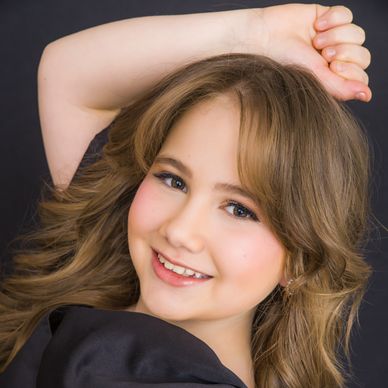 Smiling young girl with wavy hair and a black top posing against a dark background.