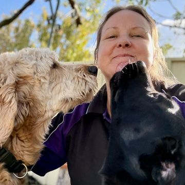 A woman enjoying affectionate licks from two dogs on a sunny day.