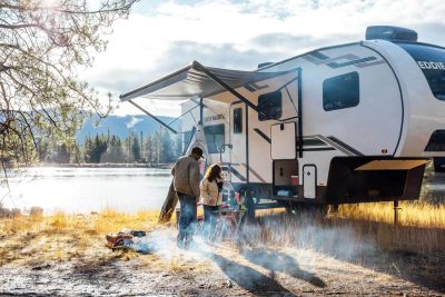 Couple camping by a lake with a trailer and campfire.