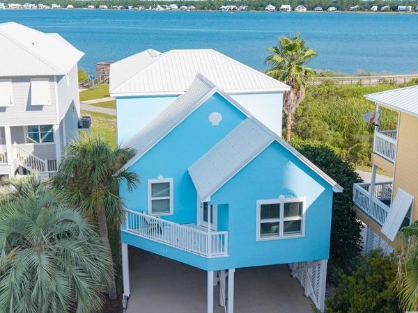 Vibrant blue stilt house near a calm waterfront with palm trees and neighboring homes.