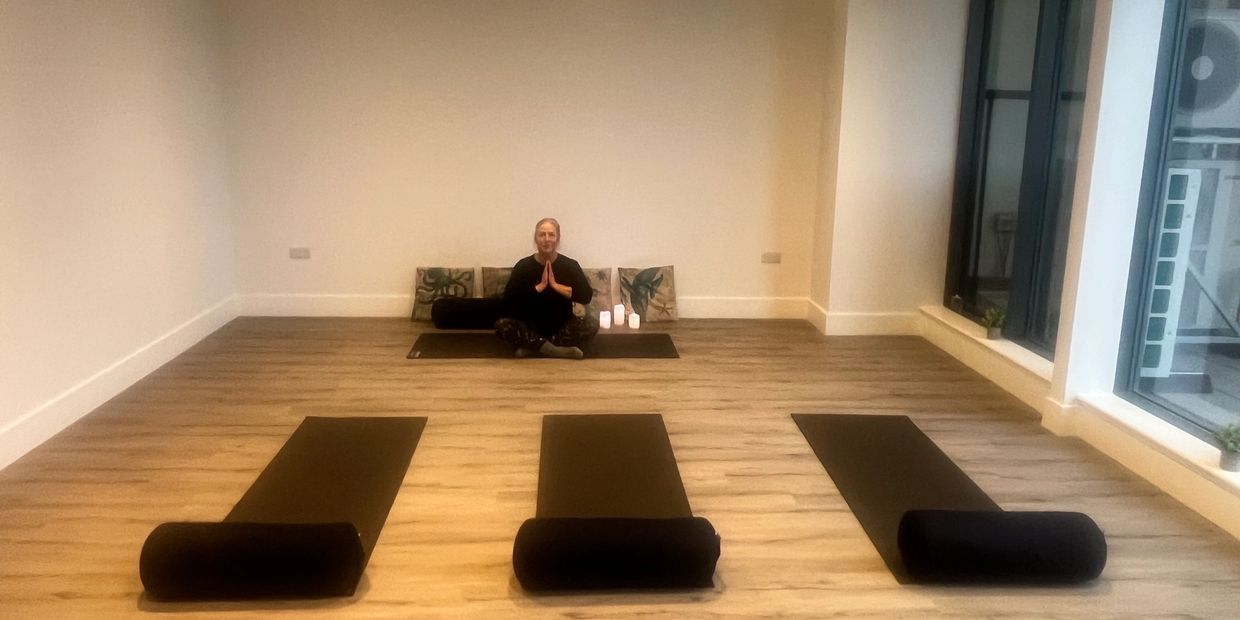 A woman sits on a yoga mat in a calm, empty studio with three mats rolled at the front.