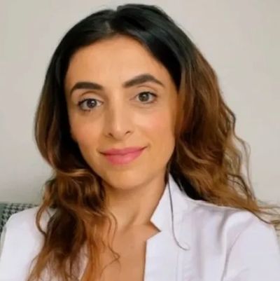 A woman with wavy hair wearing a white blazer, smiling gently.
