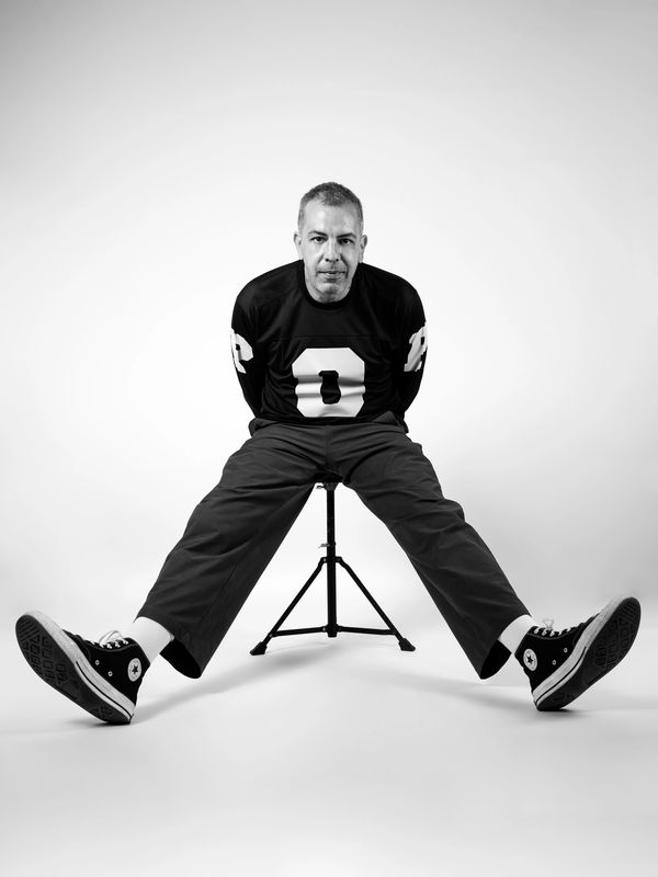 Man sitting on a stool with legs spread wide in black and white.
