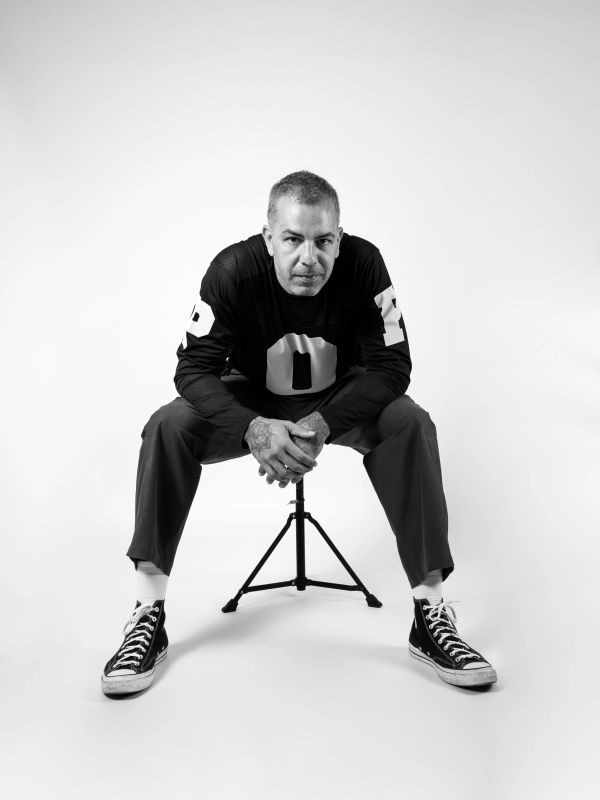 Man in casual outfit sitting on a tripod stool, looking confidently at camera.