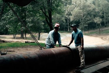 Two men working on a large metal pipe outdoors in a wooded area.