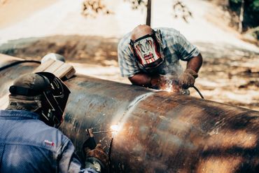 Two welders working on a large rusted metal pipe outdoors.