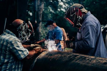 Two welders working on a large metal pipe wearing protective helmets and gloves.