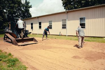 Three men leveling dirt with a tractor and rakes outside a beige building.