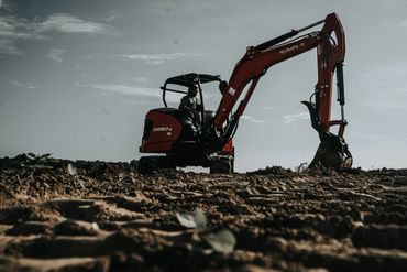 A person operating an orange Kubota excavator on a dirt field under a cloudy sky.