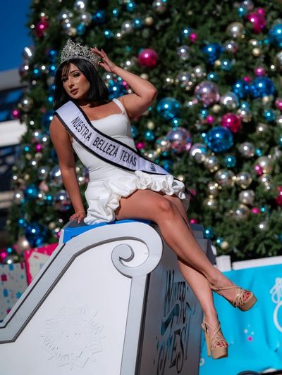 Beauty queen wearing a crown and sash, posing by a Christmas tree.