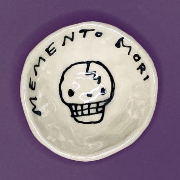 Cute smiling drawing of a skull on a small black and white ceramic dish, with the words, "MEMENTO MO