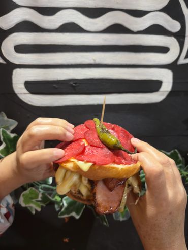 Close-up of a loaded burger with pepperoni and a green chili on top.