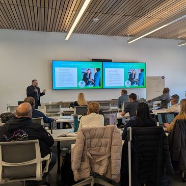 A presenter speaks to an audience in a modern conference room with dual screens.