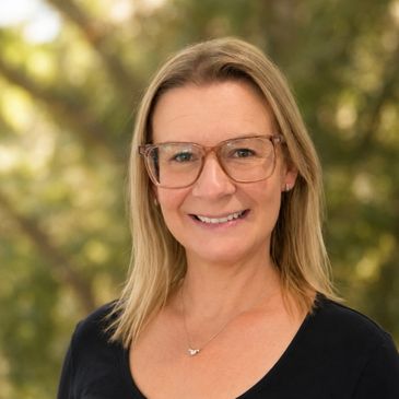 Smiling woman with glasses and blonde hair outdoors with blurred greenery background.