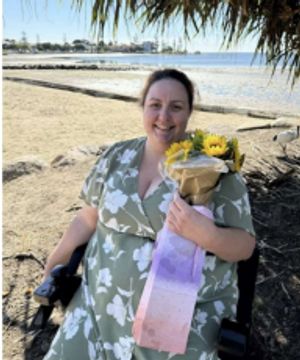Photograph of Tanya sitting in her wheelchair on the beach holding flowers.