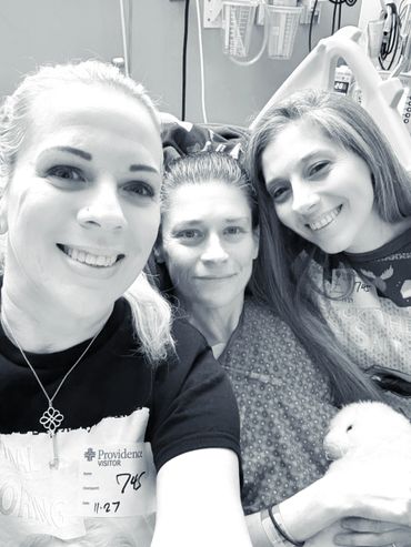 Three women smiling in a hospital room, one wearing a visitor badge.