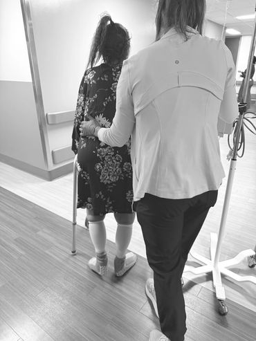 A caregiver supports a woman with a walker in a hospital hallway.
