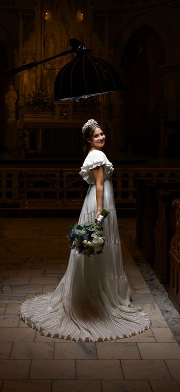 Bride in a flowing white gown holding a bouquet, lit beautifully in a dim church setting.